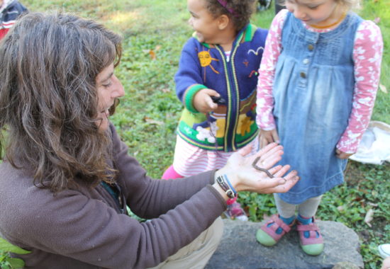 Susie Spikol holding an earthworm in her hands surrounded by several toddlers.  