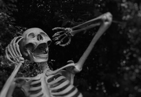 A black and white photo of a human skeleton, leaning back with its hands places up by its head.