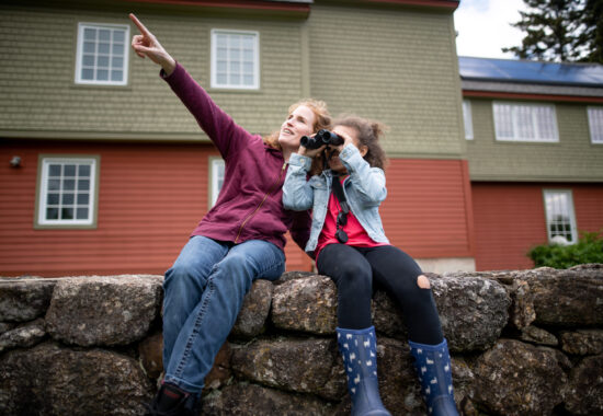 A mother and daughter sitting on a stone wall, using binoculars. 